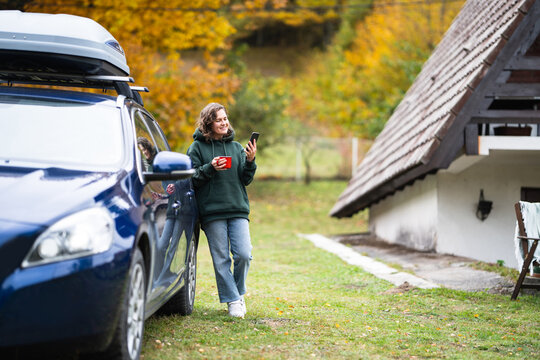 A smiling woman standing next to a blue car with a roof box, holding a red mug and using a smartphone in front of an A-frame house in autumn