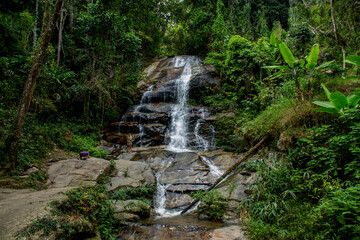 Tropical jungle waterfall in Thailand surrounded by lush green rainforest, moss-covered rocks, dense vegetation, and flowing water. The natural cascade creates a peaceful and refreshing scene © Yuliia
