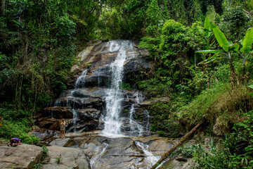 Tropical jungle waterfall in Thailand surrounded by lush green rainforest, moss-covered rocks, dense vegetation, and flowing water. The natural cascade creates a peaceful and refreshing scene © Yuliia