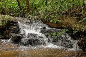 Tropical jungle waterfall in Thailand surrounded by lush green rainforest, moss-covered rocks, dense vegetation, and flowing water. The natural cascade creates a peaceful and refreshing scene © Yuliia