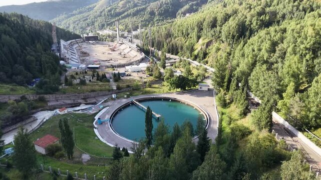ALMATY, KAZAKHSTAN - AUGUST 2026: Aerial drone flyover above circular water reservoir near Medeu mudflow control dam and high-altitude ice rink during renovation process in Medeu gorge at summer eveni