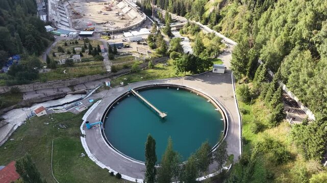 ALMATY, KAZAKHSTAN - AUGUST 2026: Aerial drone flyover above circular water reservoir near Medeu mudflow control dam and high-altitude ice rink during renovation process in Medeu gorge at summer eveni