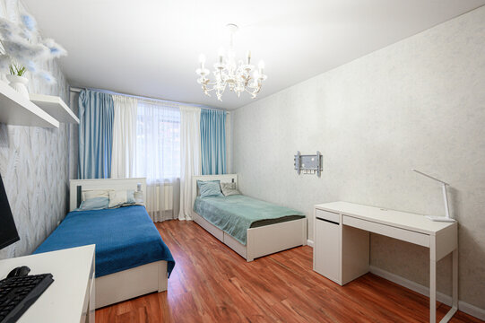 Cozy bedroom with two single beds, blue bedding, white desk, wooden floor, chandelier, blue-and-white curtains, floating shelves, minimalist decor