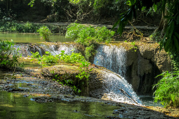Tropical jungle waterfall in Thailand surrounded by lush green rainforest, moss-covered rocks, dense vegetation, and flowing water. The natural cascade creates a peaceful and refreshing scene © Yuliia