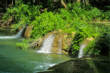 Tropical jungle waterfall in Thailand surrounded by lush green rainforest, moss-covered rocks, dense vegetation, and flowing water. The natural cascade creates a peaceful and refreshing scene © Yuliia