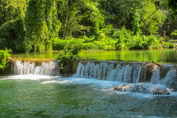 Tropical jungle waterfall in Thailand surrounded by lush green rainforest, moss-covered rocks, dense vegetation, and flowing water. The natural cascade creates a peaceful and refreshing scene © Yuliia