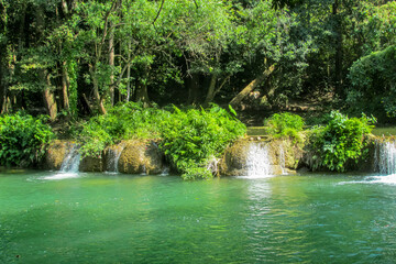 Tropical jungle waterfall in Thailand surrounded by lush green rainforest, moss-covered rocks, dense vegetation, and flowing water. The natural cascade creates a peaceful and refreshing scene © Yuliia