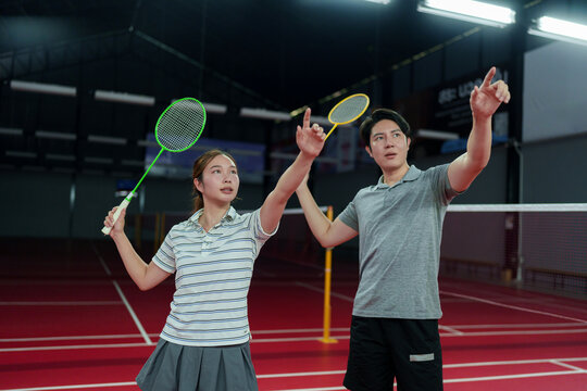 Asian coach and young woman pointing upward while holding badminton rackets on indoor court