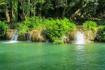 Tropical jungle waterfall in Thailand surrounded by lush green rainforest, moss-covered rocks, dense vegetation, and flowing water. The natural cascade creates a peaceful and refreshing scene © Yuliia