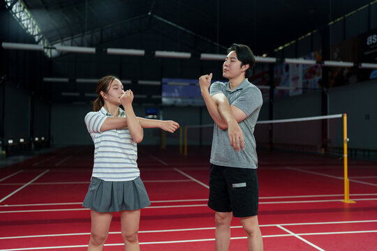 Young mixed race woman and Asian man stretching arms while badminton coach guides warmup on indoor court