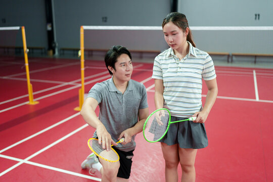 Young Asian male coach kneeling to instruct female student holding badminton racket on indoor court