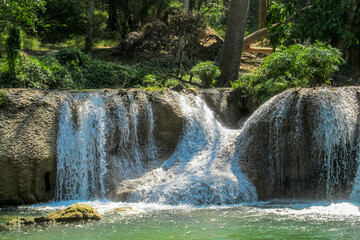Tropical jungle waterfall in Thailand surrounded by lush green rainforest, moss-covered rocks, dense vegetation, and flowing water. The natural cascade creates a peaceful and refreshing scene © Yuliia