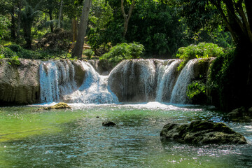 Tropical jungle waterfall in Thailand surrounded by lush green rainforest, moss-covered rocks, dense vegetation, and flowing water. The natural cascade creates a peaceful and refreshing scene © Yuliia