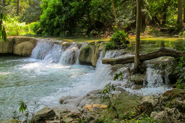 Tropical jungle waterfall in Thailand surrounded by lush green rainforest, moss-covered rocks, dense vegetation, and flowing water. The natural cascade creates a peaceful and refreshing scene © Yuliia