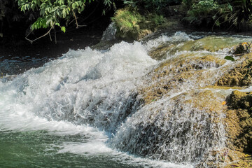 Tropical jungle waterfall in Thailand surrounded by lush green rainforest, moss-covered rocks, dense vegetation, and flowing water. The natural cascade creates a peaceful and refreshing scene © Yuliia