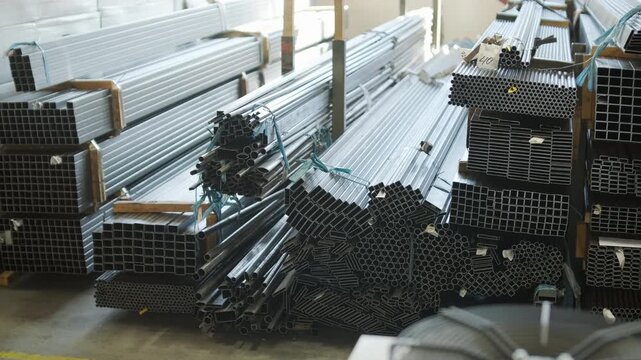Bundles of various steel and aluminum metal pipes and profiles stacked on racks in an industrial warehouse setting with a blurred foreground.