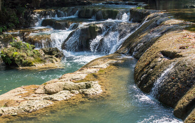 Tropical jungle waterfall in Thailand surrounded by lush green rainforest, moss-covered rocks, dense vegetation, and flowing water. The natural cascade creates a peaceful and refreshing scene © Yuliia