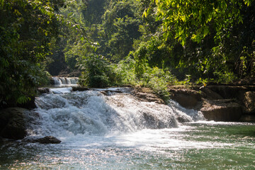 Tropical jungle waterfall in Thailand surrounded by lush green rainforest, moss-covered rocks, dense vegetation, and flowing water. The natural cascade creates a peaceful and refreshing scene © Yuliia