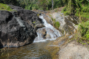 Tropical jungle waterfall in Thailand surrounded by lush green rainforest, moss-covered rocks, dense vegetation, and flowing water. The natural cascade creates a peaceful and refreshing scene © Yuliia