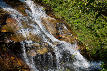 Tropical jungle waterfall in Thailand surrounded by lush green rainforest, moss-covered rocks, dense vegetation, and flowing water. The natural cascade creates a peaceful and refreshing scene © Yuliia