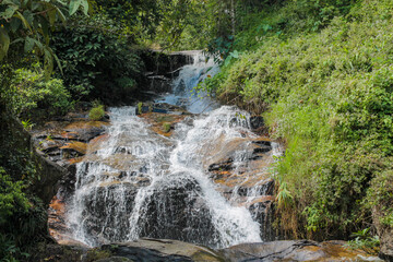 Tropical jungle waterfall in Thailand surrounded by lush green rainforest, moss-covered rocks, dense vegetation, and flowing water. The natural cascade creates a peaceful and refreshing scene © Yuliia