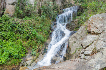 Tropical jungle waterfall in Thailand surrounded by lush green rainforest, moss-covered rocks, dense vegetation, and flowing water. The natural cascade creates a peaceful and refreshing scene © Yuliia