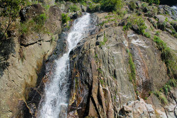 Tropical jungle waterfall in Thailand surrounded by lush green rainforest, moss-covered rocks, dense vegetation, and flowing water. The natural cascade creates a peaceful and refreshing scene © Yuliia