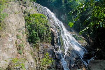 Tropical jungle waterfall in Thailand surrounded by lush green rainforest, moss-covered rocks, dense vegetation, and flowing water. The natural cascade creates a peaceful and refreshing scene © Yuliia