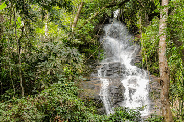 Tropical jungle waterfall in Thailand surrounded by lush green rainforest, moss-covered rocks, dense vegetation, and flowing water. The natural cascade creates a peaceful and refreshing scene © Yuliia