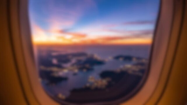 Aerial view of a coastal landscape at sunset seen through an airplane window. The soft focus creates a dreamy atmosphere over the dark shoreline and glowing city lights below.