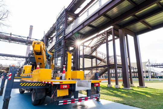A bright yellow aerial work truck parks firmly on flat ground inside an old industrial zone.