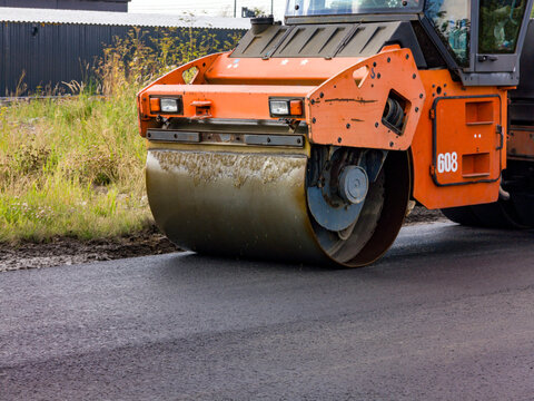 Road roller compacts fresh asphalt on a construction site. Heavy machinery levels a paved surface beside grass and industrial fencing.