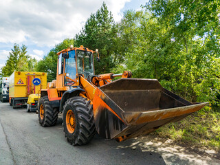 Fototapeta premium Front loader with bucket stands beside a utility truck on a wooded roadside. Heavy equipment and warning signs appear beneath a summer sky.