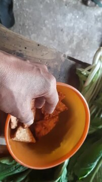 A top-down view showing a hand picking up a piece of crispy fried tempeh from an orange bowl. Tempeh is a traditional Indonesian fermented soybean food and a source of plant-based (vegan) protein.