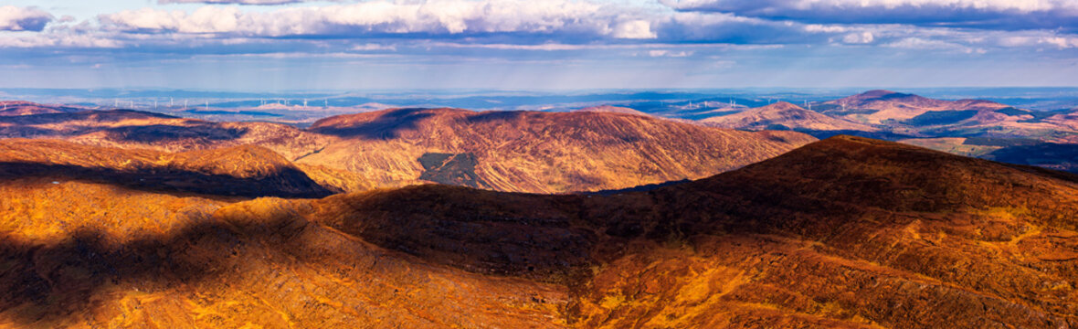 A panoramic view shows rolling hills covered in vegetation, with shadows cast across the landscape under a cloudy sky.