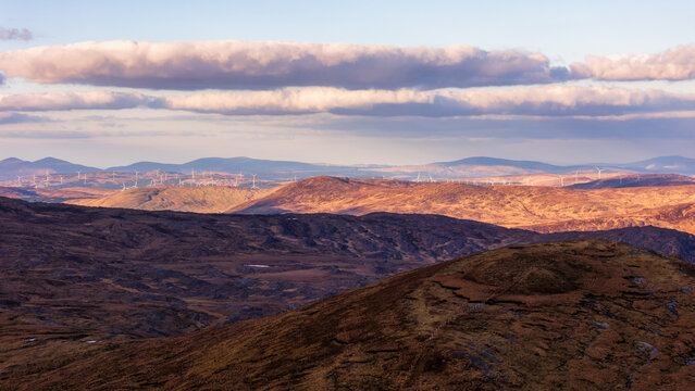 Golden hour light bathes the rolling mountains of Cork, as sunset shadows stretch across the landscape and distant turbines turn.