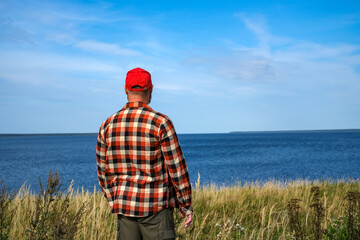 man seen from behind, wearing a red cap and plaid shirt, stands among tall grass by the sea and looks at the horizon. Concept of freedom, travel, peaceful solitude and connection with nature. © Dmitrii Labrador
