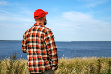man seen from behind, wearing a red cap and plaid shirt, stands among tall grass by the sea and looks at the horizon. Concept of freedom, travel, peaceful solitude and connection with nature. © Dmitrii Labrador