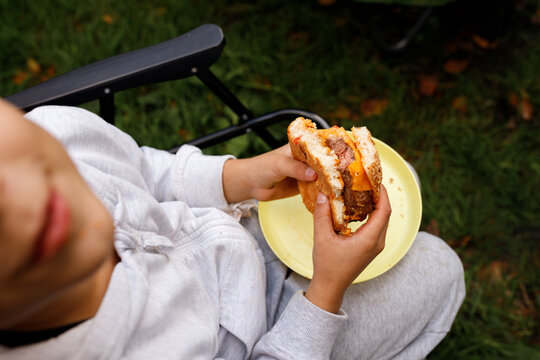 Boy eating homemade burger close up hands holding bitten cheeseburger over plate child sitting in camping chair family picnic outdoor cookout barbecue background lawn backyard leisure lifestyle