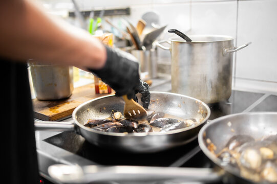 A chef wearing black gloves stirs fresh mussels in a stainless steel frying pan on a stove in a professional restaurant kitchen, preparing a seafood dish.