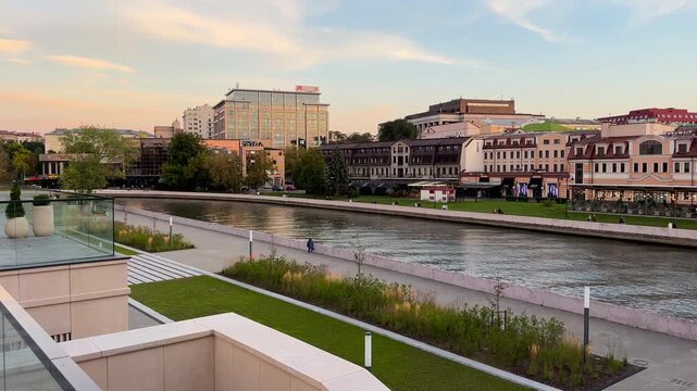 Riverbank with buildings and park on sunset in city. Svisloch River embankment. Landscaped waterfront with pedestrian and historic architecture of city center. Belarus, Minsk, September 16, 2026