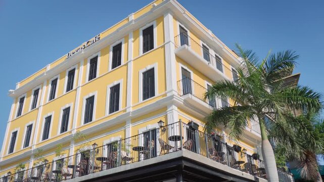 Historic yellow coffee shop building with empty terrace tables facing Plaza Hijas de Tampico under clear blue sky framed by tropical palm trees