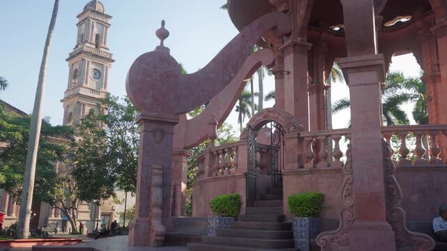 Pink stone octopus kiosk in Tampico main square with majestic cathedral facade standing under bright sunrise sky surrounded by tropical palm trees