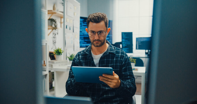 Young Caucasian male developer uses digital tablet to inspect software coding algorithms at office desk. Programming development, Artificial intelligence machine learning, Secure database concept.