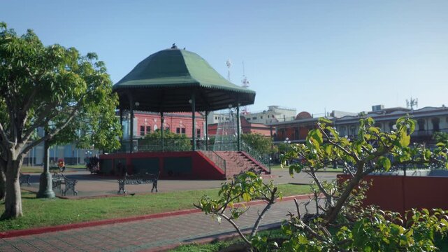 Historic green iron gazebo in Plaza de la Libertad framed by tropical plants during sunrise with pedestrians walking past classic nineteenth century French style buildings in Tampico