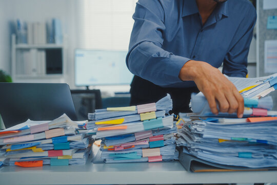 Closeup overworked young Latin businessman reading document in large stack of paperwork file at office desk. Tax audit deadline, accounting stress, corporate burnout, bookkeeping, bureaucracy concept.