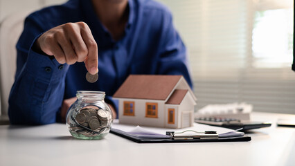 A person places a coin into a savings jar beside a model house. The image represents saving for a home purchase, mortgage down payment, housing expenses, property insurance and financial planning. © DK_STUDIO