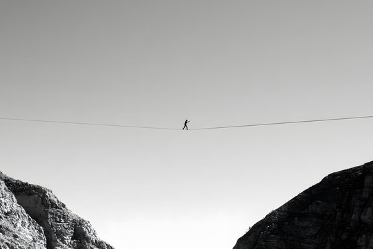 Silhouette of a person high-lining between two high cliffs against a clear sky, emphasizing balance and danger. A stark black and white image of an extreme sport.