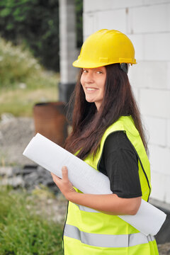 Young female construction worker in hi vis vest, helmet and gloves, carrying cable roll on her shoulder, smiling, bare new house wall near
