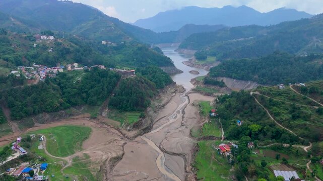 Aerial view of a flood-devastated river corridor covered in thick sediment deposits and mudflats cutting through rural Nepalese foothills
